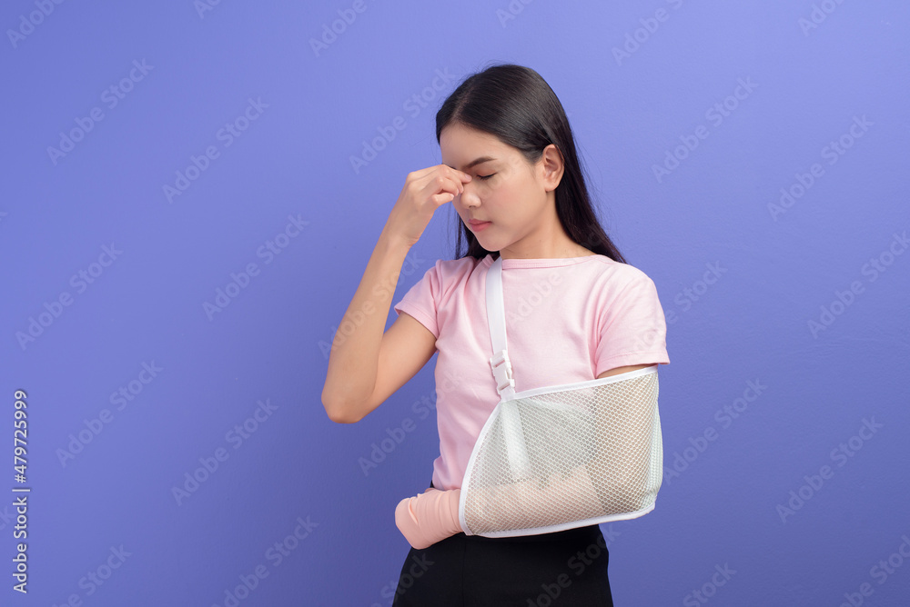 Portrait of young woman stressed and frustrated with an injured arm in a sling over blue background in studio, insurance and healthcare concept Portrait of young woman stressed and frustrated with an injured arm in a sling over blue background in studio, insurance and healthcare concept