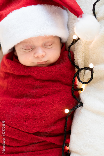 Top View Portrait First Days Of Life Newborn Cute Funny Sleeping Child Baby In Santa Hat Wrapped In Red Diaper At White Garland Background. Merry Christmas, Happy New Year, Infant, Childhood, Winter