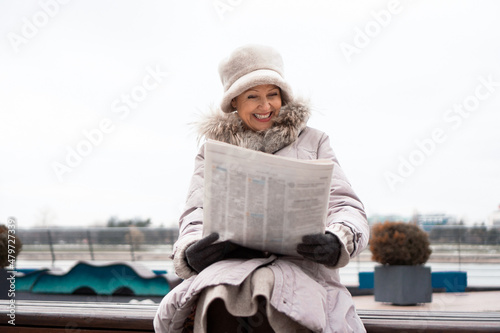 A beautiful, elegant senior woman sits by the river and reads the newspaper on a cold winter day.  She is wearing a long winter jacket with fur around the neck and a warm hat and gloves. photo