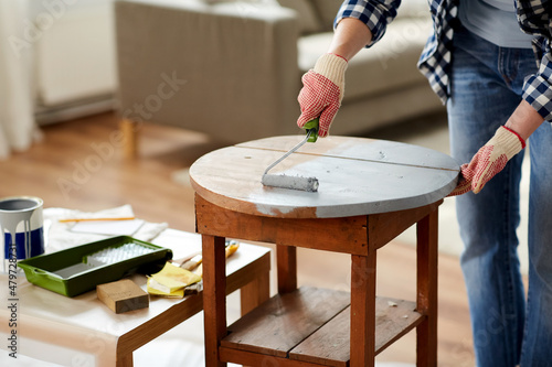 furniture renovation, diy and home improvement concept - close up of woman in gloves with paint roller painting old wooden table in grey color photo