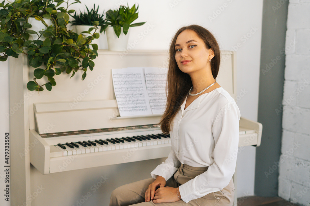 Portrait of smiling young woman pianist looking at camera sitting at classical white piano in classroom. Successful attractive female musician performer preparing for classes at home studio. Portrait of smiling young woman pianist looking at camera sitting at classical white piano in classroom. Successful attractive female musician performer preparing for classes at home studio.