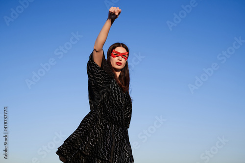 Portrait of strong supergirl in black dress and red face mask making fist pump, protesting for freedom and equality on blue sky background фототапет