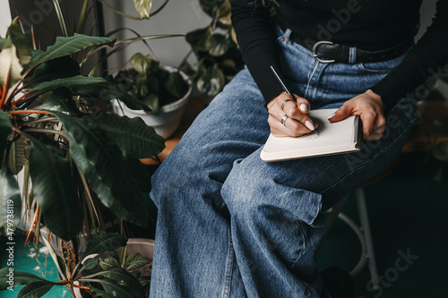 Woman is writing something in her notebook. She is sitting in a cozy cafe surrounded by plants.