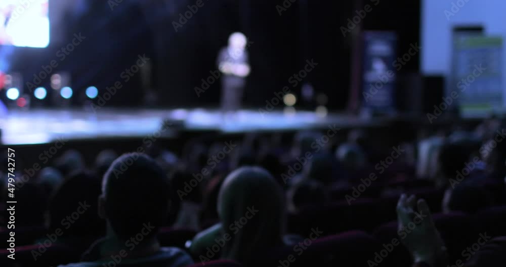 Back view of an audience sitting at a business conference. A camera is moving from the back of the audience to the stage, showing the speaker giving the presentation. Conference and training concept.