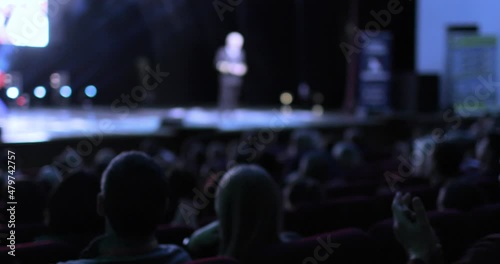 Back view of an audience sitting at a business conference. A camera is moving from the back of the audience to the stage, showing the speaker giving the presentation. Conference and training concept.