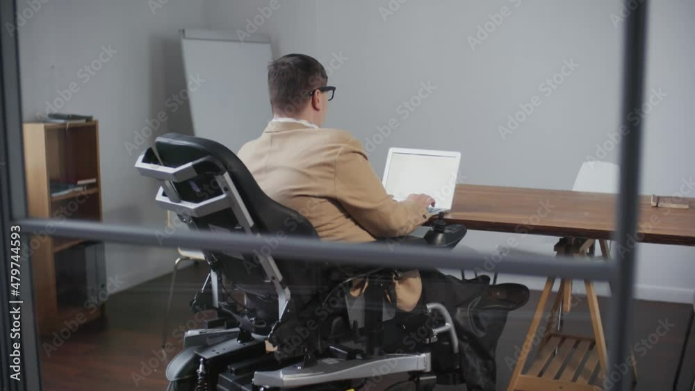 Rear view of intelligent man with disabilities dressed formally, sitting at desk in office at daytime, using portable computer Rear view of intelligent man with disabilities dressed formally, sitting at desk in office at daytime, using portable computer