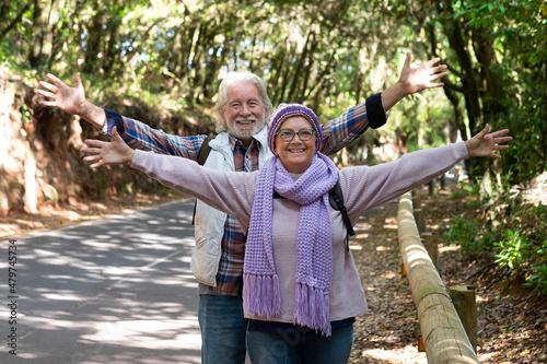 Senior couple of hikers standing with open arms in the shadow of the woods enjoying the freedom and healthy lifestyle. Active happy couple of retired smiling looking at camera
