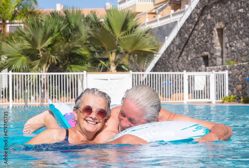 Cheerful mature adult active happy senior couple of grandparents relaxing in the swimming pool floating on mattress outdoors in water. Smiling retired people enjoying vacation healthy lifestyle