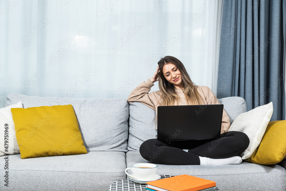 Young happy woman sitting on sofa at home drinking coffee or tea watching the movie on her laptop computer. Carefree female enjoying her free time with hot drink and online dating on the internet. Young happy woman sitting on sofa at home drinking coffee or tea watching the movie on her laptop computer. Carefree female enjoying her free time with hot drink and online dating on the internet.