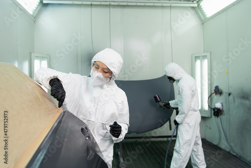 Workwoman in goggles and protective suit working with car part near colleague with aerograph in garage. photo