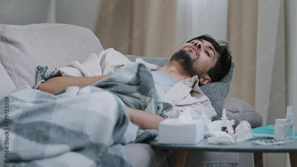 Sick health man arabic spaniard guy feeling symptoms of coronavirus covid coughing flu suffering from illness infectious virus lying on couch under blanket at home looking at table with medications