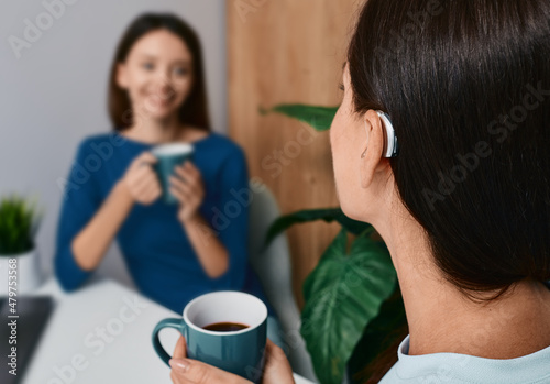 Adult woman with a hearing impairment uses a hearing aid to communicate with her female friend during tea drinking at home. Hearing solutions photo