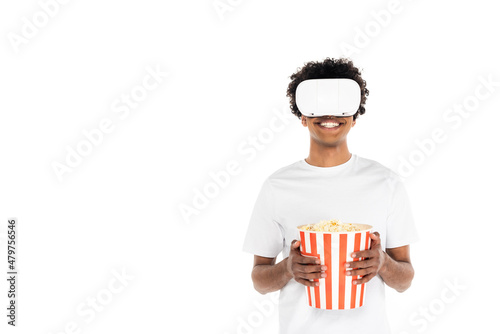 cheerful african american man with big bucket of popcorn gaming in vr headset isolated on white.