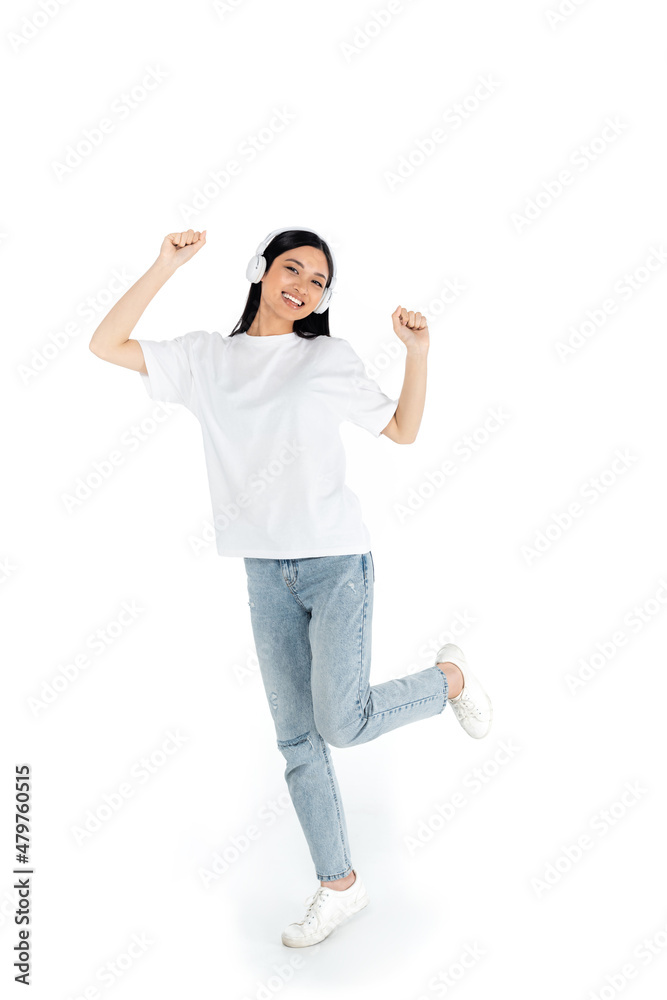 full length view of pleased asian woman in jeans and headphones dancing on white. full length view of pleased asian woman in jeans and headphones dancing on white.