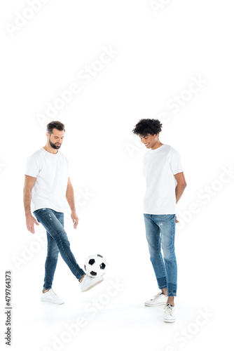 smiling african american man standing with hands behind back near friend playing with soccer ball on white.