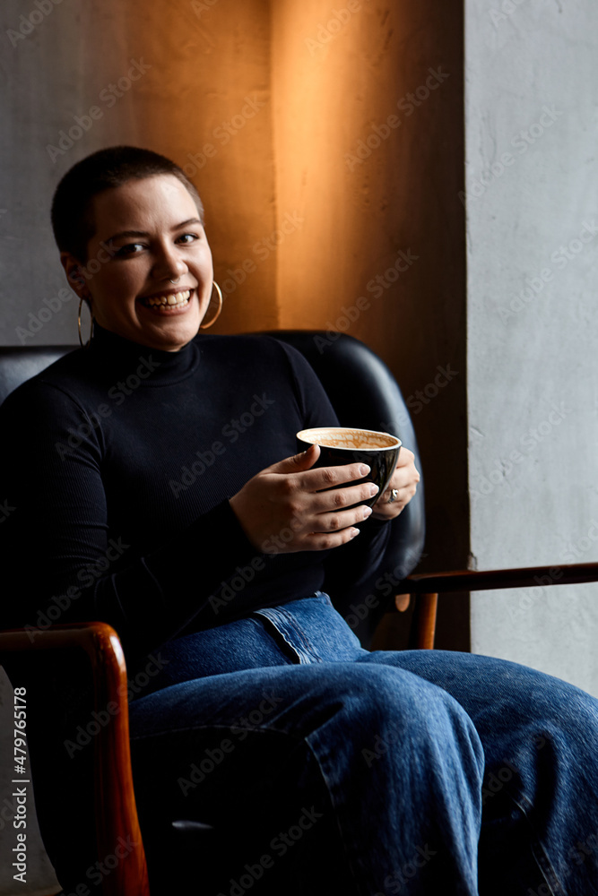 A young girl with glasses, with short hair and a nose piercing is sitting in a cafe, drinks coffee.