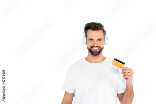 pleased man in headphones looking at camera while holding credit card isolated on white.