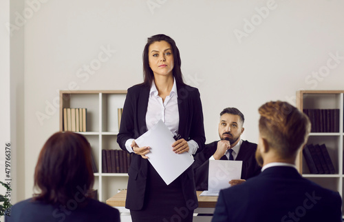 Portrait of female lawyer or attorney who represents plaintiff in lawsuit during court trial. Serious unsmiling woman holding documents standing in courtroom after her speech in front of audience