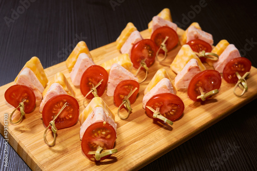 Canapes on bamboo skewers made of homemade ham, pieces of cheese in the shape of triangles, cherry tomatoes on a wooden board.