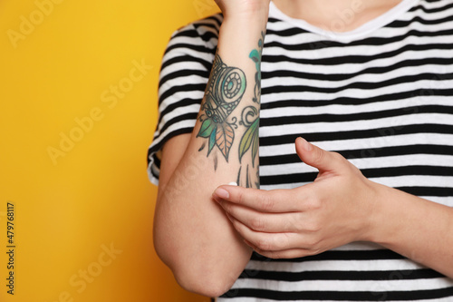 Woman applying cream on her arm with tattoos against yellow background, closeup. Space for text