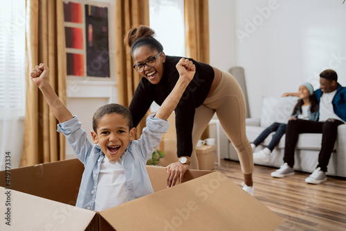 Caring mom playing with baby, moving cardboard box across living room floor with sweet son sitting on it, joy of moving into new apartment, smiling little boy putting hands up from good time