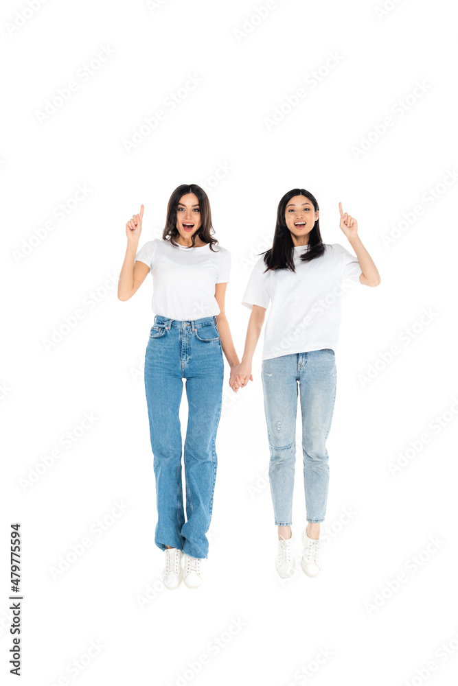 full length view of cheerful multiethnic women pointing with fingers while levitating isolated on white. full length view of cheerful multiethnic women pointing with fingers while levitating isolated on white.