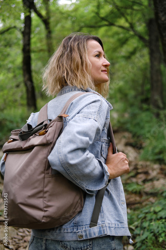 A young Caucasian woman walks along a forest mountain road. Casual blue denim clothes and backpack. Travel, healthy lifestyle and freedom.