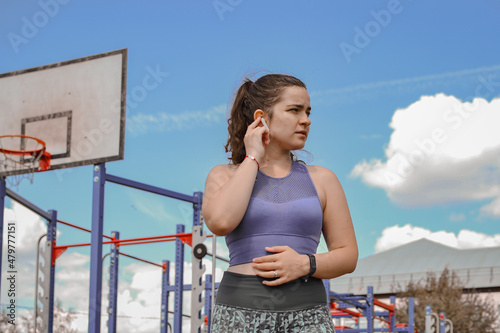 A young woman on the background of the sky and the sports field performed a set of fitness exercises rests, switches music and listens to a voice message sent by work colleagues, looks away.