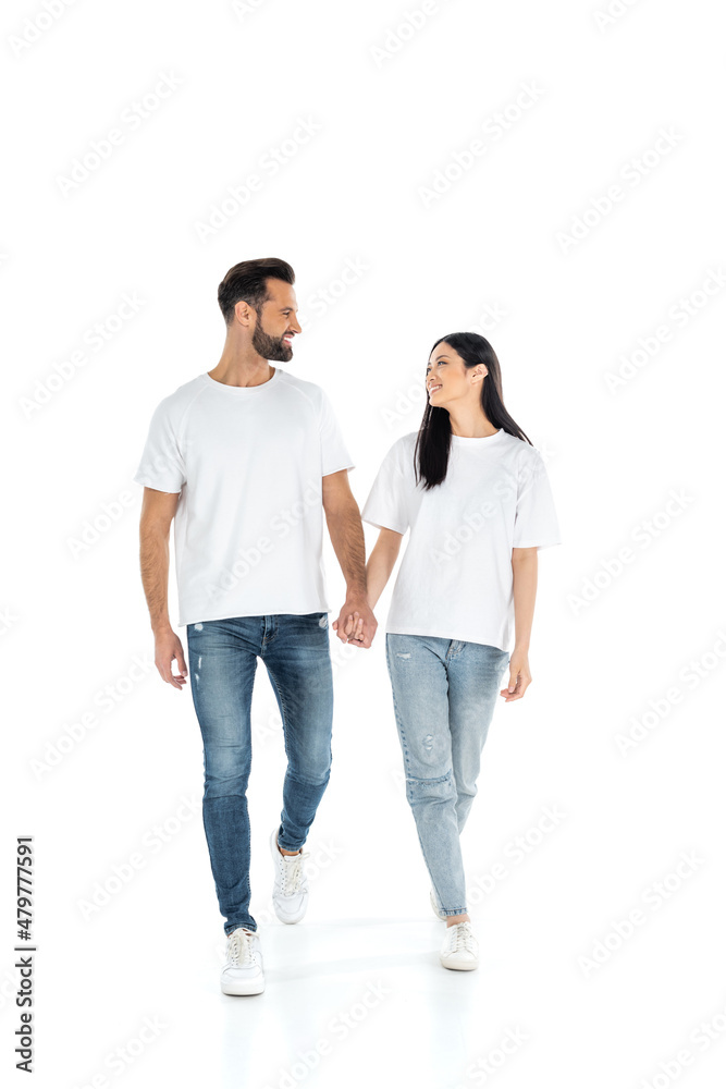 full length view of happy interracial couple looking at each other and holding hands while walking on white.