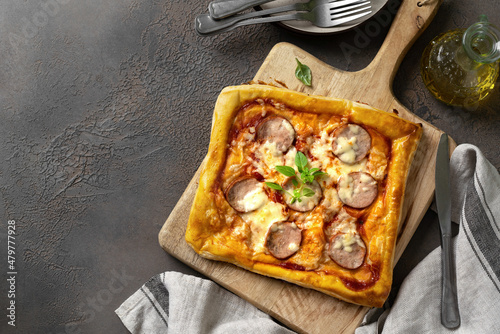 Homemade puff pastry pizza with salami, mozzarella, tomatoes and basil on the kitchen table. Delicious layered pie with sausage and cheese on a dark culinary background top view with a copyspace