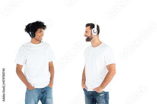 interracial men in headphones standing with hands in pockets of jeans and looking at each other isolated on white.