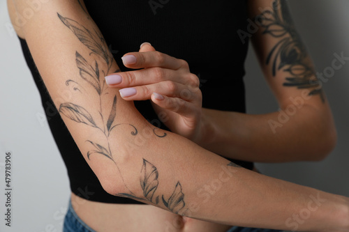 Woman applying cream on her arm with tattoos against light background, closeup photo