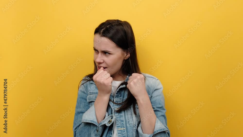 Portrait young caucasian woman 20s stands with clenched fists and furious look, ready to punch, expressing aggression, wears blue denim jacket, posing isolated over yellow color background in studio Portrait young caucasian woman 20s stands with clenched fists and furious look, ready to punch, expressing aggression, wears blue denim jacket, posing isolated over yellow color background in studio