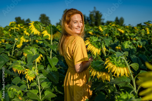 Sweet smiling young red-haired caucasian woman in a yellow dress looks into the camera, winked in a sunflower field on a sunny day