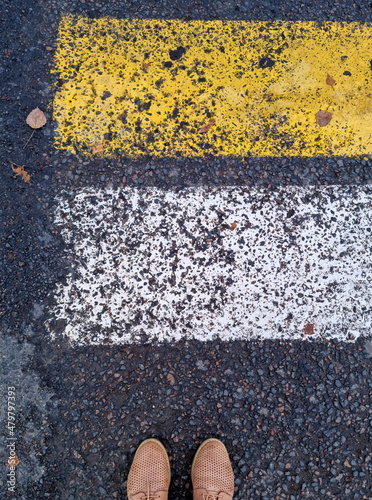 A person in yellow shoes stands in front of a pedestrian crossing, top view. Rough texture of asphalt, yellow and white road markings on the road, zebra crossing.