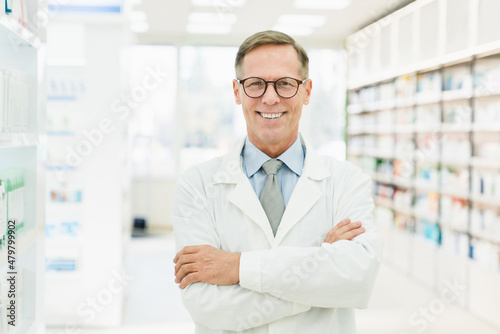 Smiling male caucasian middle-aged mature confident pharmacist druggist in white medical coat standing with arms crossed in pharmacy drugstore