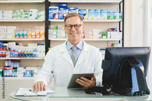 Successful smiling male caucasian middle-aged mature chemist druggist pharmacist holding digital tablet for checking medicines standing at cash point desk in pharmacy drugstore