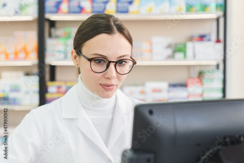 Young caucasian female chemist pharmacist druggist working on computer, looking at screen searching drugs, medicines, pills for customers in drugstore pharmacy
