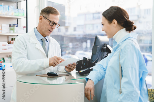 Male caucasian middle-aged mature pharmacist looking at medical prescription selling drugs medications medicines to young female customer buyer in pharmacy