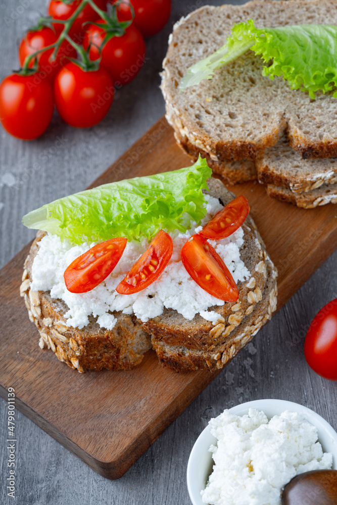 Slices of rye bread with cottage cheese and tomatoes on wooden plate on grey background