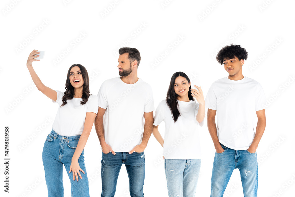 skeptical men standing with hands in pockets near happy interracial women taking selfie isolated on white. skeptical men standing with hands in pockets near happy interracial women taking selfie isolated on white.