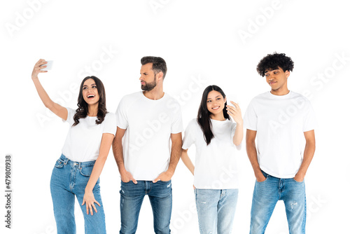 skeptical men standing with hands in pockets near happy interracial women taking selfie isolated on white.