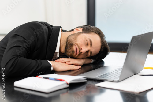 Tired young businessman in formal wear taking a nap on the workplace, male office employee in suit sleeping on the desk in office, takes a break, overworked or feels boredom