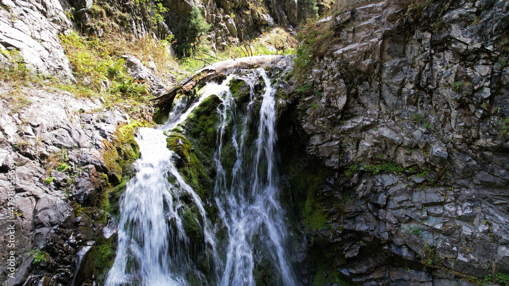 A mountain waterfall in a rocky gorge. The view from the drone is of steep cliffs, moss, green plants and the river. There are stones lying. Splashes from the water. There is a log lying. Almaty