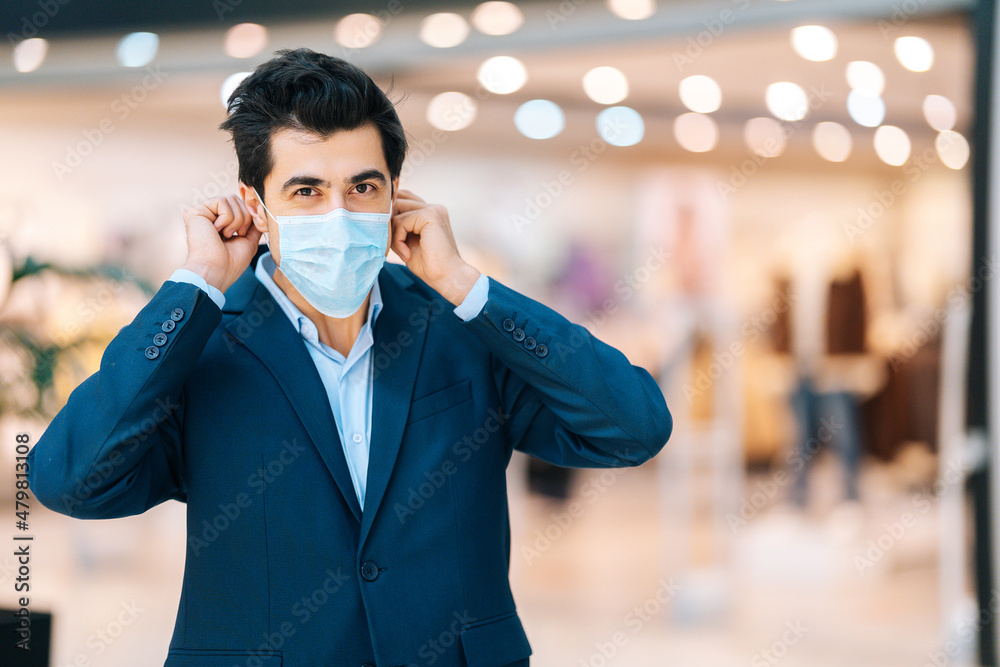 Portrait of handsome young man in stylish suit wearing protection face mask looking at camera in hall of mall centre, blurred background. Front view of confident male applying mask.
