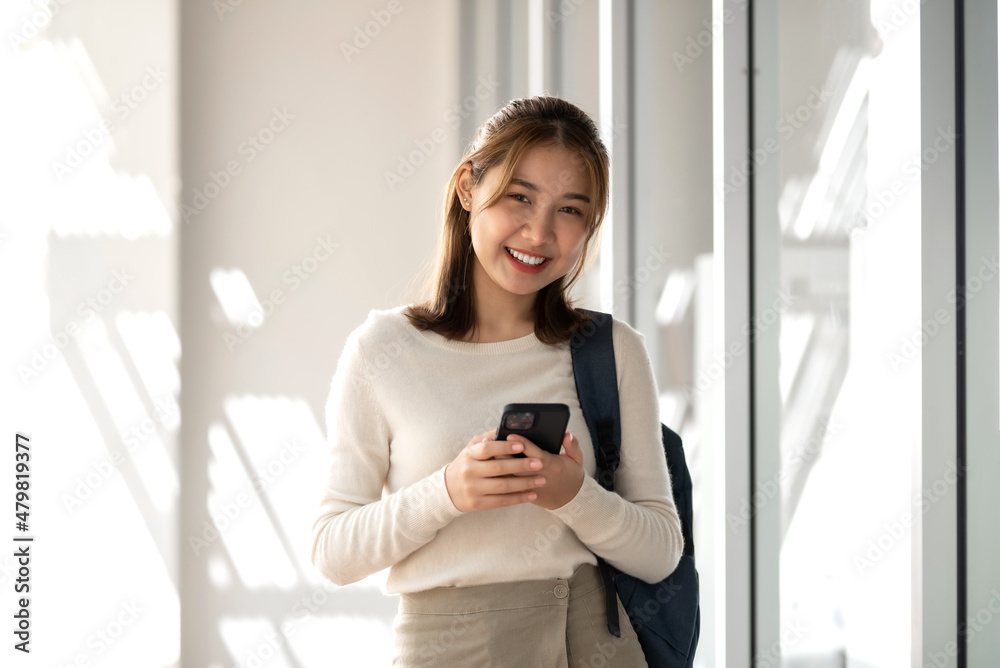 Pretty smiling happy young Asian student standing outside the classroom hand holding smartphone.  Looking at camera.