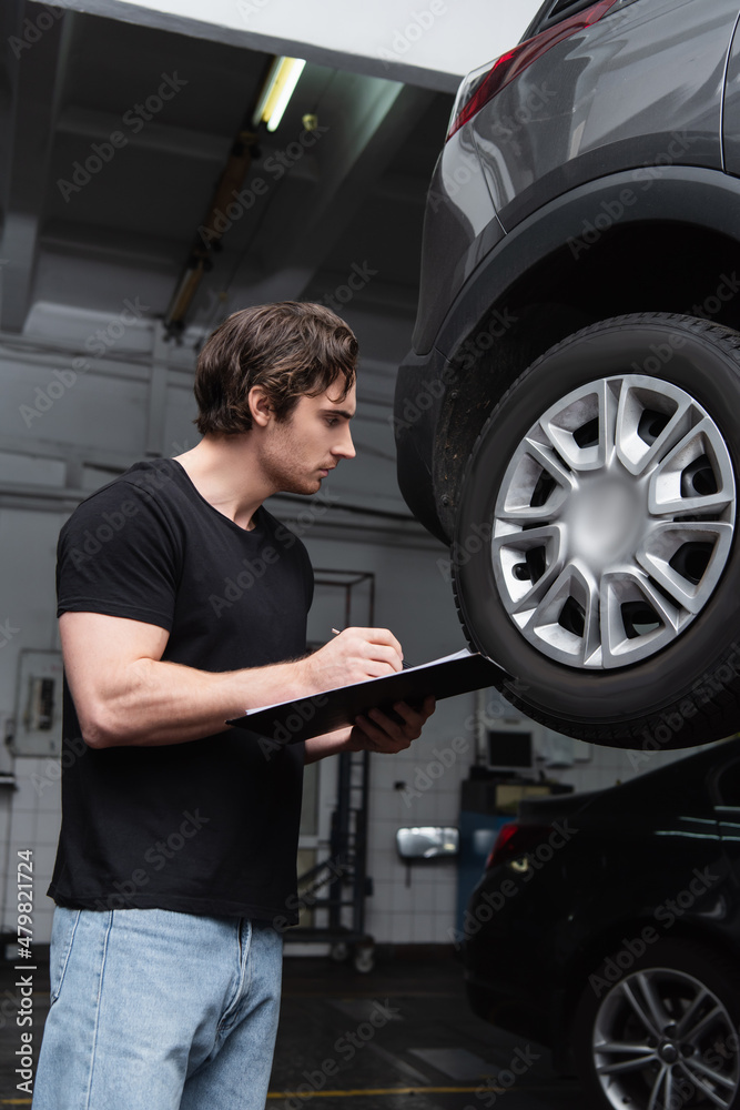 Side view of mechanic writing on clipboard near car wheel in garage.