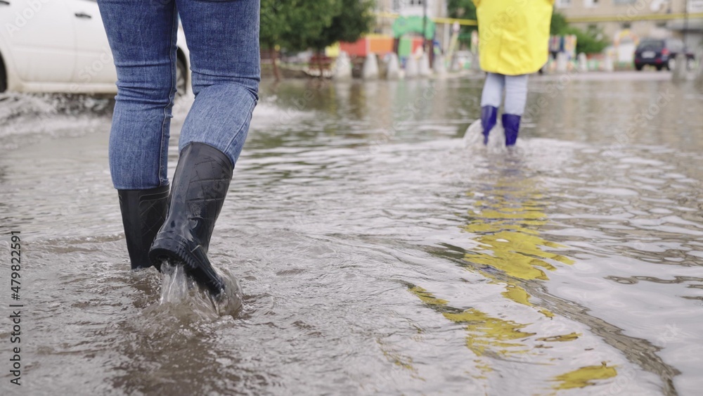 two people walk through puddles of flooded city rubber boots on their feet, teamwork, girls summer raincoats, lot of rainwater is poured on street, natural weather phenomena, walking boots on road