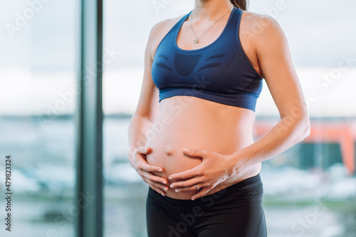 Free Download Unrecognisable pregnant woman in sportswear with chain and pendant on neck caressing belly on window background. - GFXLoad