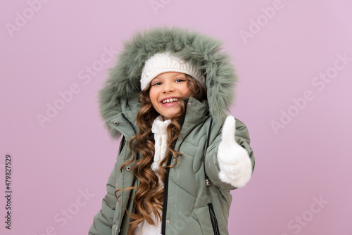 a little girl in a winter jacket and a warm knitted hat gives a thumbs up. a child in winter clothes on an isolated background.